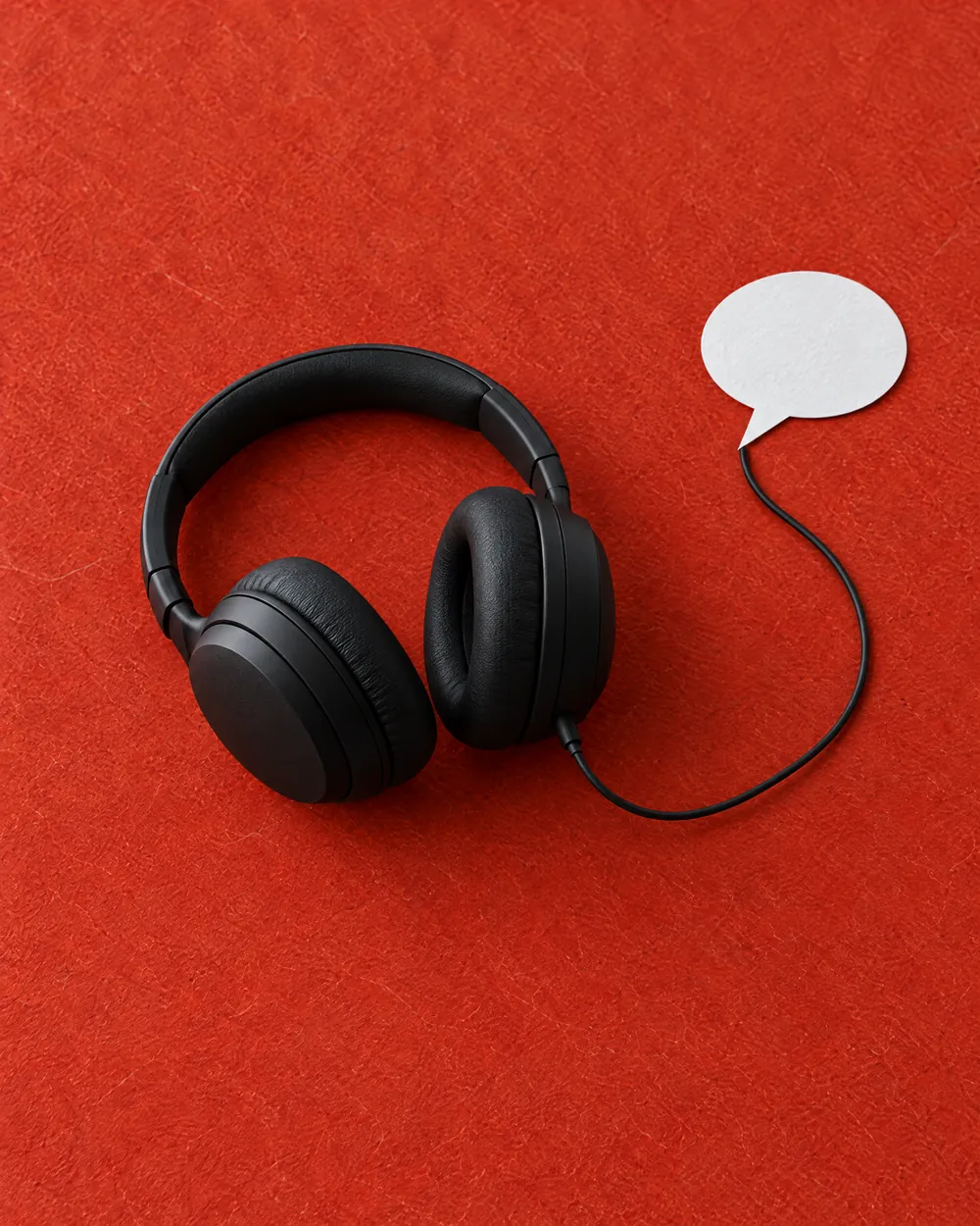 Large black headphones whose single cable leads to a flat speech bubble cut from white paper, on vermilion washi.